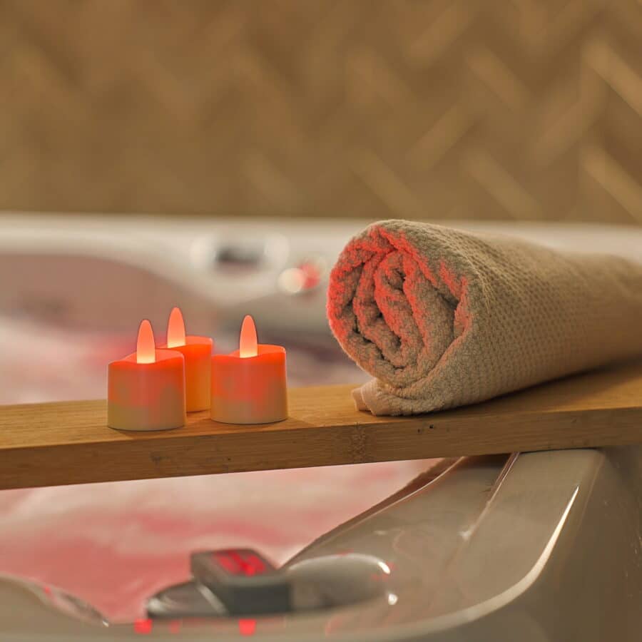Een opgerolde beige handdoek en drie verlichte waterdichte RGB oplaadbare waxinelichtjes met afstandsbediening (68 uur brandtijd) staan op een houten plank bij een jacuzzi, voor een ontspannen spa-ervaring.