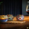 Drie Theelichthouder 9 CM Dorsetto kleurrijke kraltjes kandelaars met brandende kaarsen staan op een geweven mat op een houten tafel, naast een glas rode wijn, en creëren een zacht verlichte en gezellige sfeer.