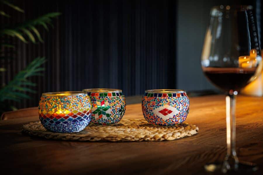 Drie Theelichthouder 9 CM Dorsetto kleurrijke kraltjes kandelaars met brandende kaarsen staan op een geweven mat op een houten tafel, naast een glas rode wijn, en creëren een zacht verlichte en gezellige sfeer.