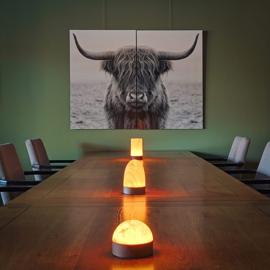Een houten eettafel met drie gloeiende zoutlampen en een witte, marmeren, oplaadbare LED-tafellamp met timer in houtlook staat tussen de stoelen, onder een zwart-witfoto van een hooglandkoe op een groene muur.