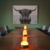 Een houten vergadertafel met drie witte, goudkleurige oplaadbare LED tafellampen met timerfuncties wordt omringd door grijze stoelen. Aan de muur hangt een zwart-wit portret in twee panelen van een langhoornige Highland koe.