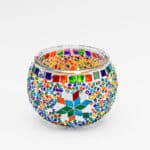 De Waxinelichthouder 9 CM Dorsetto Rainbown is een ronde glazen waxinelichthouder met kleurrijke mozaïekpatronen, sterrendesigns en veelkleurige kralen tegen een effen witte achtergrond.