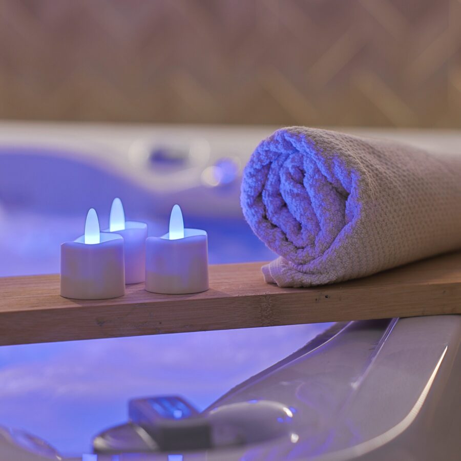 Een opgerolde handdoek rust op een houten plank boven een bubbelende jacuzzi, met 12 oplaadbare RGB led theelichtjes (waterdicht, met afstandsbediening en timerfunctie) die een ontspannen spasfeer creëren.