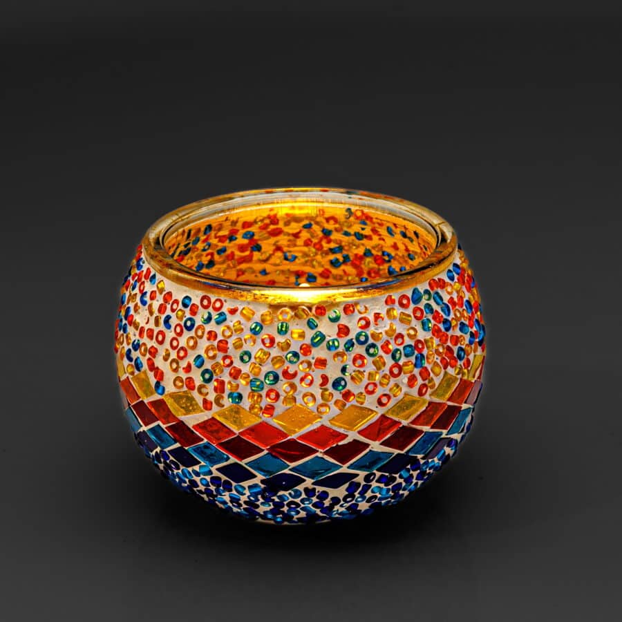 De Waxinelichthouder 9 cm 3 colormix is een ronde glazen mozaïekhouder met levendige blauwe, rode, gele en oranje kralen en tegeltjes op een donkere achtergrond. De binnenkant gloeit warm wanneer hij wordt aangestoken.