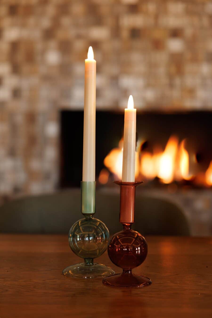 Twee warmwitte, oplaadbare dinerkaarsen (set van 12, 270 mm, brandtijd 130 uur, afstandsbediening) gloeien op een houten tafel, hun licht weerkaatst in kleurrijke glazen houders met een open haard zacht vervaagd op de achtergrond.