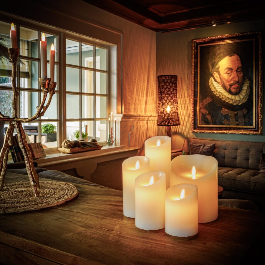 Een gezellige woonkamer met Oplaadbare led kaars (30 uur brandtijd, afstandsbediening, 7,5 x 12,5 cm) gloeiend op een houten tafel, een kandelaar, grijze bank, groot portret van een man in antieke kleding, en warm licht van een geweven vloerlamp.