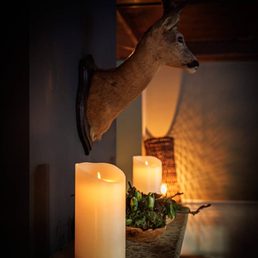 Een opgezette hertenkop hangt boven een mantel aan een donkere muur, geflankeerd door twee Oplaadbare led kaarsen (30 uur brandtijd, afstandsbediening, 7,5x10 cm) en decoratief groen in een gezellige, schemerig verlichte kamer.