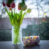 Een glazen vaas met kleurrijke tulpen en twee Theelichthouder 9 CM Dorsetto kleurrijke kraltjes kandelaars rusten op een donker oppervlak, met een vage achtergrond van bomen en daken.