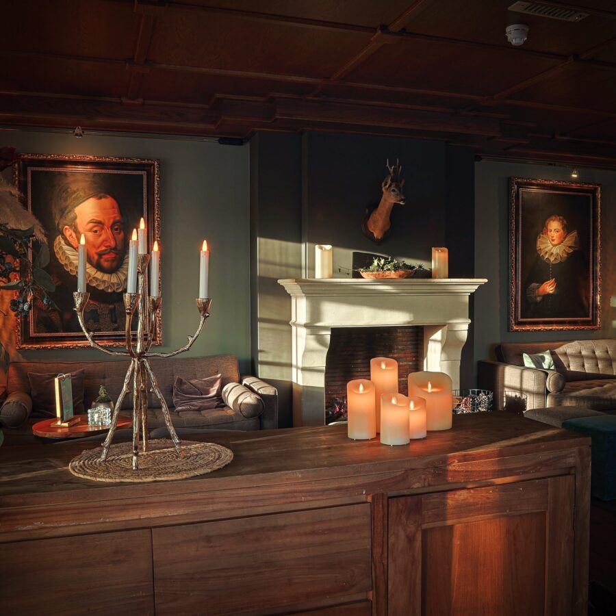 Een gezellige, schemerig verlichte kamer heeft een houten tafel met Oplaadbare led kaarsen (7,5x12,5cm, 30 uur brandtijd, afstandsbediening) en een marmeren open haard versierd met pilaarkaarsen, die een warme vintage sfeer creëren.