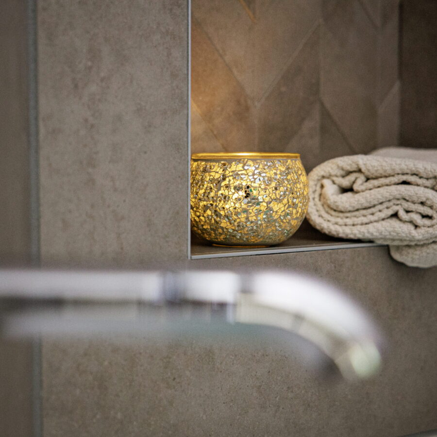 Een Theelichthouder zwart 9 CM DORSETTO met een gloeiende kaars rust op een plank naast een netjes opgevouwen handdoek in een moderne badkamer met grijs betegelde muren.