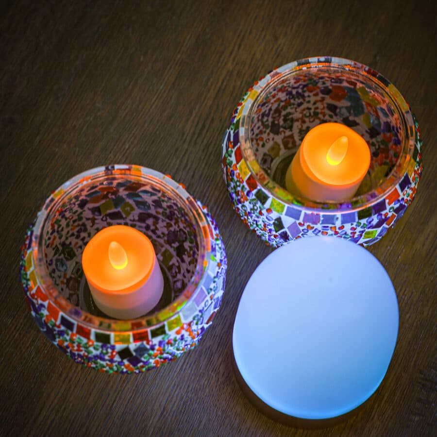Twee Theelichthouder 9 CM Dorsetto kleurrijke kaarsenhouders met LED-kaarsen erin staan op een houten oppervlak naast een gloeiend wit ovaalvormig object.