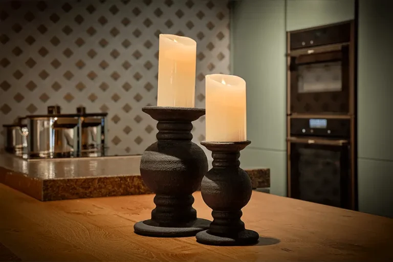 Twee brandende pilaarkaarsen of led waxinelichtjes op donkere houders staan op een houten aanrecht. Op de achtergrond staan roestvrijstalen potten, een wand met patroon en ingebouwde ovens, die een warme, gezellige sfeer creëren.