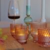 Een houten gedekte tafel met brandende roze glazen kandelaars, 12 oplaadbare waxinelichtjes (175 uur brandtijd, waterdicht, met afstandsbediening en timerfunctie), een groene kandelaar met een witte kaars, een wijnglas met rode wijn en wat groene druiven.