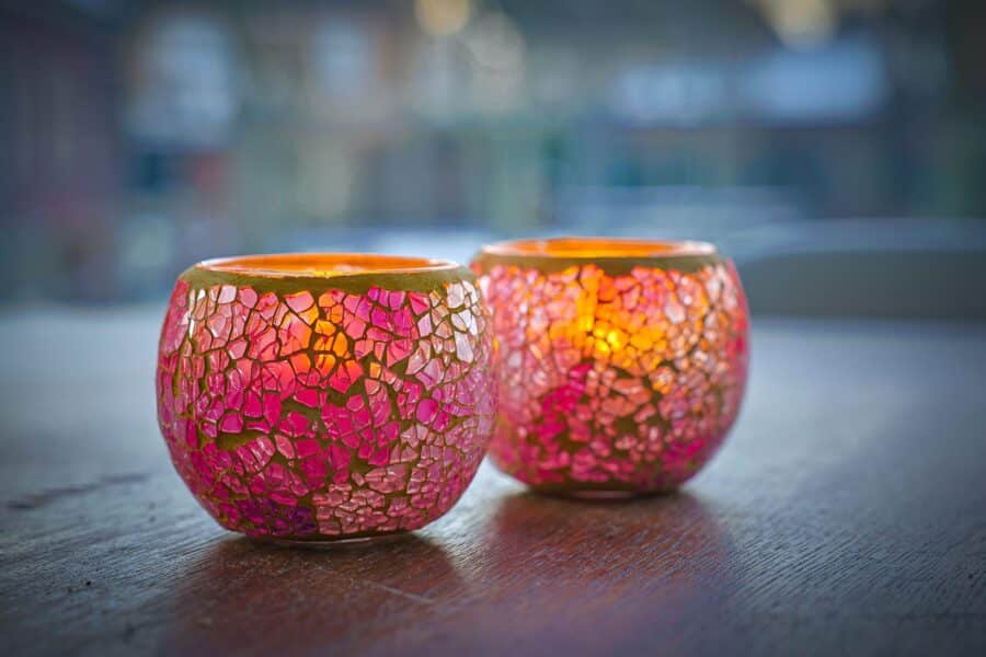 Twee Waxinelichthouder 8,5 CM Dorsetto mozaïek roos kandelaars met roze en goud mozaïek glas zitten op een houten oppervlak, elk warm opgloeiend van een brandende kaars. De zacht vervaagde achtergrond draagt bij aan de gezellige sfeer.