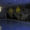 Een binnenzwembad onder een plafond verlicht door de LED Sterrenhemel RGB (180 sterren) met afstandsbediening, met een muurschildering van een mediterrane kust en een wit standbeeld bij de muur.