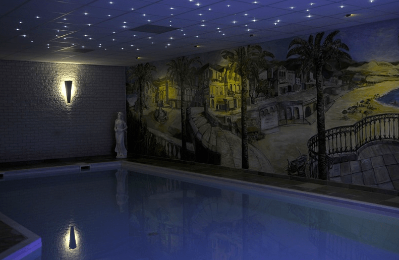 Een binnenzwembad onder een plafond verlicht door de led sterrenhemel rgb (180 sterren) met afstandsbediening, met een muurschildering van een mediterrane kust en een wit standbeeld bij de muur.