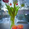 Een glazen vaas met tulpen en twee Waxinelichthouder 8,5 CM Dorsetto mozaïek roos kandelaars staan op een donkere tafel, met een winters buitengezicht en huizen zichtbaar in de wazige achtergrond.