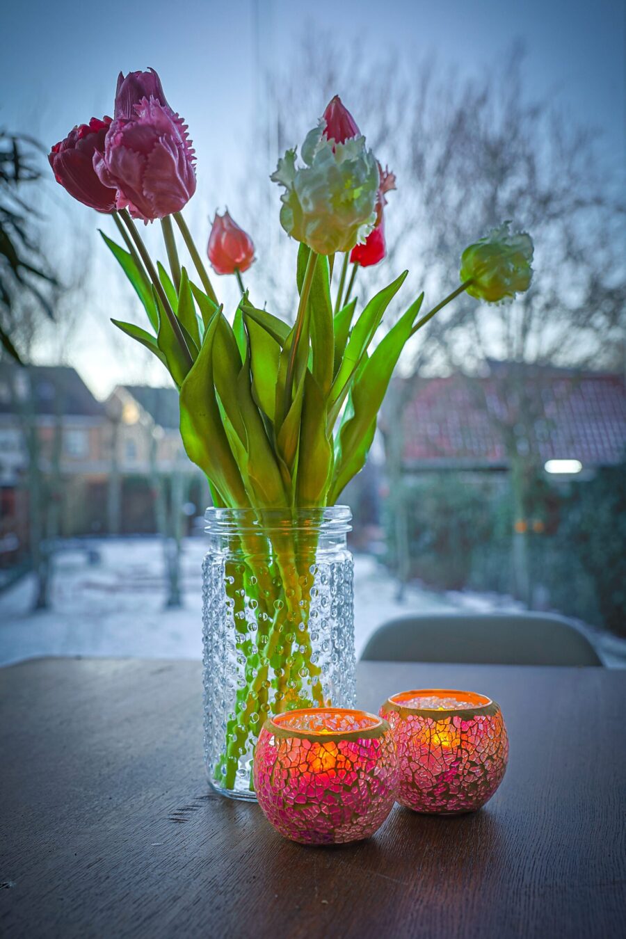 Een glazen vaas met tulpen en twee Waxinelichthouder 8,5 CM Dorsetto mozaïek roos kandelaars staan op een donkere tafel, met een winters buitengezicht en huizen zichtbaar in de wazige achtergrond.