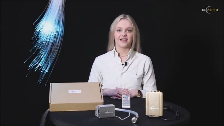 Een vrouw zit aan een tafel met een LED Sterrenhemel RGB (180 sterren) met afstandsbediening kit-bruine doos, apparaten, afstandsbediening, kabels. Blauwe glasvezelstrengen gloeien aan de linkerkant; het logo van Dorsetto verschijnt in de rechterbovenhoek.