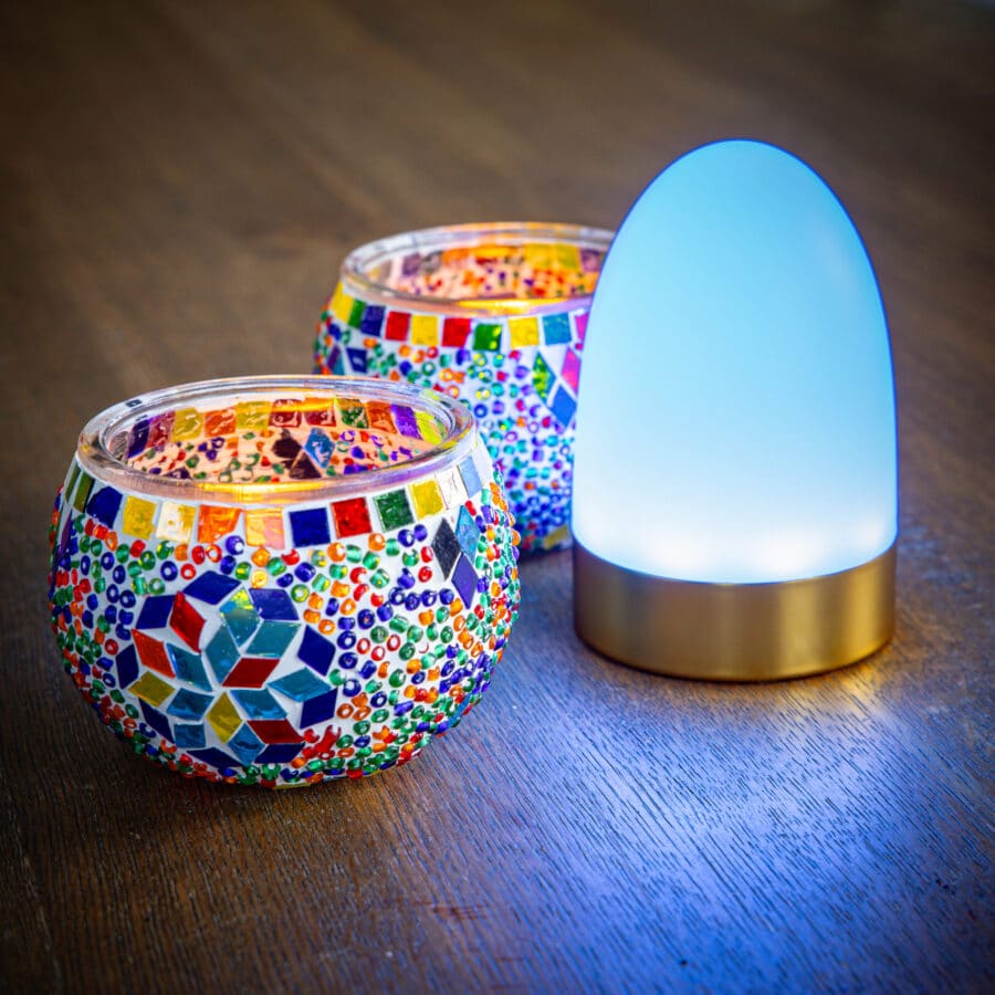 Twee Theelichthouder 9 CM Dorsetto kleurrijke kraaltjes met levendige patronen staan naast een moderne, blauwgloeiende lamp op een houten oppervlak.