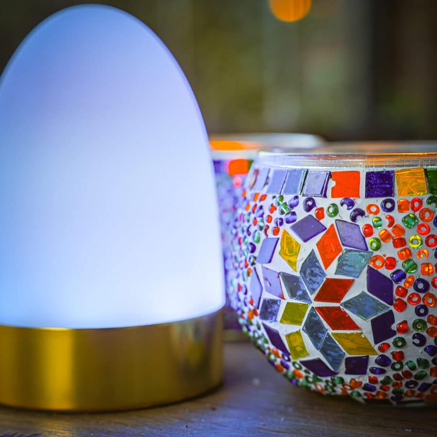 Een stralend witte lamp met een gouden voet staat naast de Theelichthouder 9 CM Dorsetto kleurrijke kraltjes, waarvan de levendige patronen en kralen charme toevoegen aan het houten oppervlak.