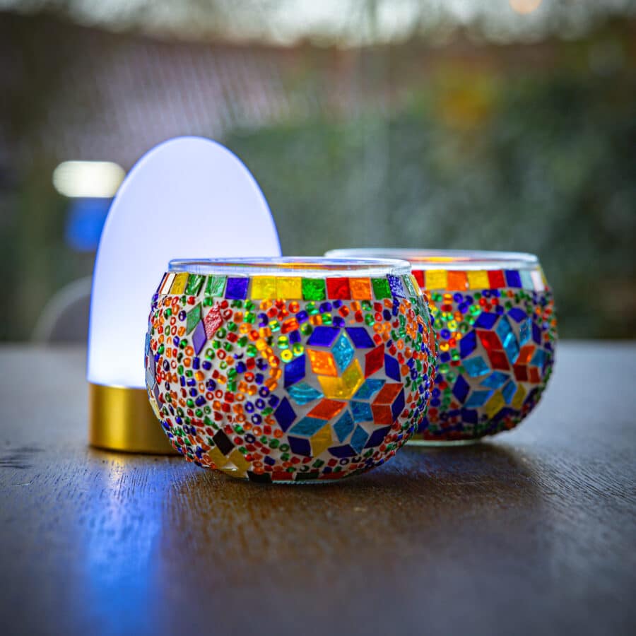 De Theelichthouder 9 CM Dorsetto kleurrijke kraltjes mozaïek glazen houder en een kleine moderne LED lamp rusten op een houten oppervlak, tegen een vage buitenachtergrond.