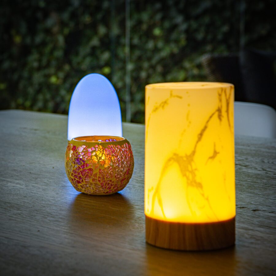 Een Waxinelichthouder 8,5 CM Dorsetto mozaïek roos gloeit met een mozaïekpatroon naast twee andere decoratieve lampen op een houten tafel, tegen een bladerrijke achtergrond.