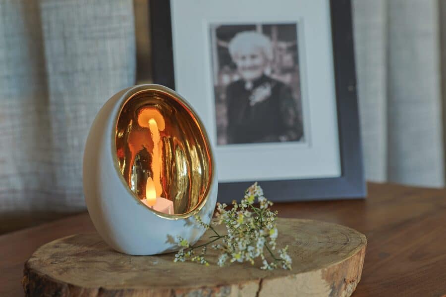 Een kandelaar met een gouden binnenkant bevat 12 oplaadbare waxinelichtjes (175 uur brandtijd, waterdicht, met afstandsbediening en timerfunctie) en staat op een houten schijf bij witte bloemen; een onscherpe foto staat op de achtergrond.