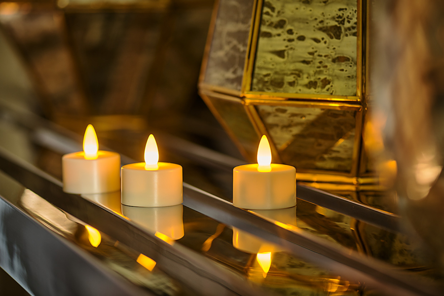 Drie kleine, oplaadbare waxinelichtjes met warmgeel licht zijn op een rij geplaatst op een reflecterend oppervlak, met gouden decoratieve objecten zacht vervaagd op de achtergrond.