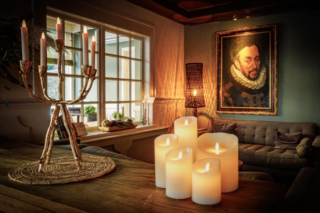 Een gezellige woonkamer met led-kaarsen die gloeien op een houten tafel, een kandelaar, een bank, een groot raam en een portret van een man in historische kleding aan de warm verlichte muur.