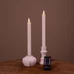 Een set van twee witte led dinerkaarsen 2026 (400 uur brandduur, oplaadbaar) in keramische houders staat op een houten oppervlak met een kleine zwarte afstandsbediening ervoor. De effen bruine achtergrond maakt het tafereel compleet.