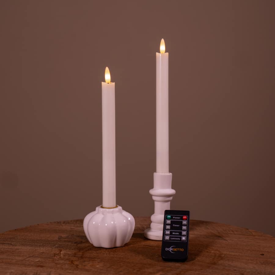 Een set van twee witte led dinerkaarsen 2026 (400 uur brandduur, oplaadbaar) in keramische houders staat op een houten oppervlak met een kleine zwarte afstandsbediening ervoor. De effen bruine achtergrond maakt het tafereel compleet.