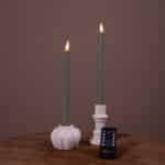 Twee verlichte Dinerkaarsen LED 400 uur brandduur oplaadbaar wit (set van 2) in witte keramische houders staan op een houten oppervlak, met een kleine zwarte afstandsbediening ervoor. De achtergrond is effen en gedempt bruin.