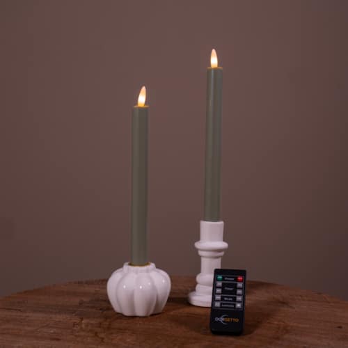 Twee verlichte Dinerkaarsen LED 400 uur brandduur oplaadbaar wit (set van 2) in witte keramische houders staan op een houten oppervlak, met een kleine zwarte afstandsbediening ervoor. De achtergrond is effen en gedempt bruin.