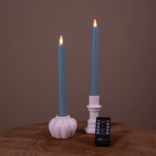 Twee witte Dinerkaarsen LED kaarsen (400h oplaadbaar, set van 2) met kunstmatige vlammen zitten op een houten oppervlak. Aan de voorkant staat een kleine zwarte Dogetto afstandsbediening. De achtergrond is effen bruin.