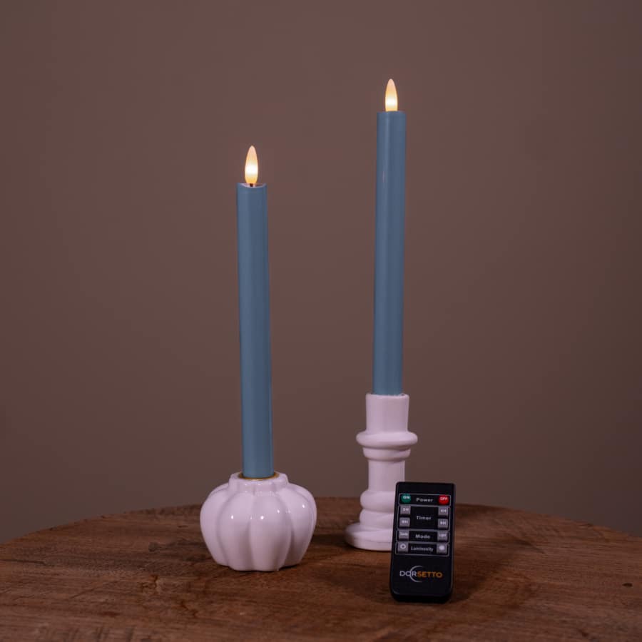 Twee witte Dinerkaarsen LED kaarsen (400h oplaadbaar, set van 2) met kunstmatige vlammen zitten op een houten oppervlak. Aan de voorkant staat een kleine zwarte Dogetto afstandsbediening. De achtergrond is effen bruin.