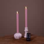 Twee witte Dinerkaarsen LED kaarsen (set van 2, 400 uur oplaadbaar) met realistische vlammen zitten in houders op een houten oppervlak. Een kleine zwarte afstandsbediening ligt ervoor. De achtergrond is effen bruin.