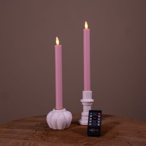 Twee witte Dinerkaarsen LED kaarsen (set van 2, 400 uur oplaadbaar) met realistische vlammen zitten in houders op een houten oppervlak. Een kleine zwarte afstandsbediening ligt ervoor. De achtergrond is effen bruin.