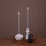 Twee witte led dinerkaarsen LED-kaarsen (set van 2), met een brandtijd van 400 uur en oplaadbare functie, staan in houders op een houten oppervlak. Een afstandsbediening ligt ervoor, alles tegen een effen bruine achtergrond.