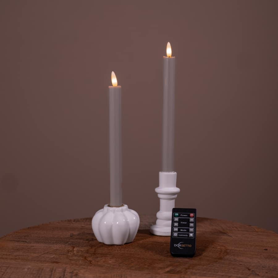 Twee witte led dinerkaarsen LED-kaarsen (set van 2), met een brandtijd van 400 uur en oplaadbare functie, staan in houders op een houten oppervlak. Een afstandsbediening ligt ervoor, alles tegen een effen bruine achtergrond.