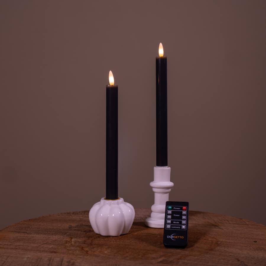 Twee witte Dinerkaarsen LED-kaarsen (set van 2, oplaadbaar, brandtijd 400 uur) met nepvlammen in houders staan op een houten oppervlak. Een kleine afstandsbediening ligt ervoor, tegen een effen bruine achtergrond.