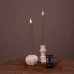 Twee witte oplaadbare LED dinerkaarsen (set van 2, 400 uur) met vlammen staan in houders op een ronde houten tafel, met een kleine zwarte "DESETTO" afstandsbediening ervoor. De achtergrond is effen bruin.