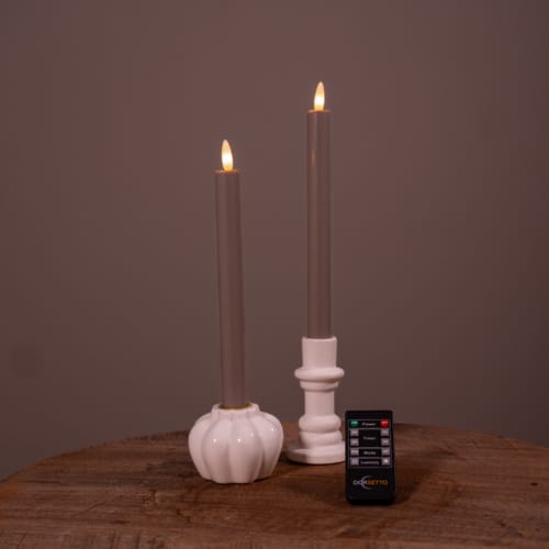 Twee witte oplaadbare LED dinerkaarsen (set van 2, 400 uur) met vlammen staan in houders op een ronde houten tafel, met een kleine zwarte "DESETTO" afstandsbediening ervoor. De achtergrond is effen bruin.