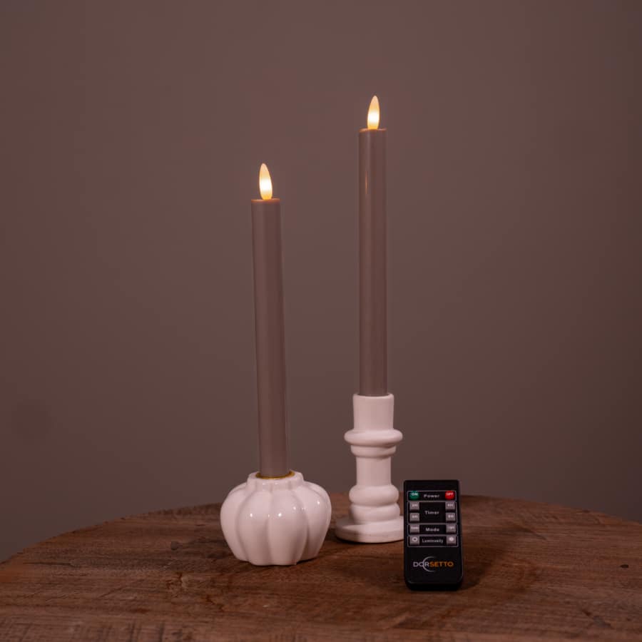 Twee witte oplaadbare LED dinerkaarsen (set van 2, 400 uur) met vlammen staan in houders op een ronde houten tafel, met een kleine zwarte "DESETTO" afstandsbediening ervoor. De achtergrond is effen bruin.