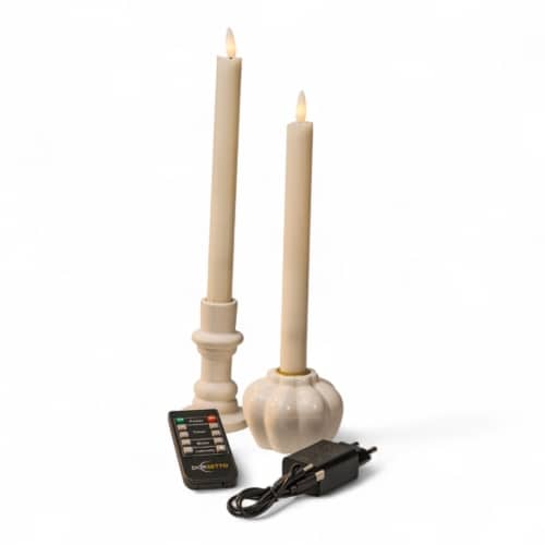 Twee crèmekleurige LED conische kaarsen in witte houders staan rechtop. Voor hen liggen een zwarte afstandsbediening met het label Deligetto en een zwarte stroomadapter met een snoer, alles tegen een witte achtergrond.
