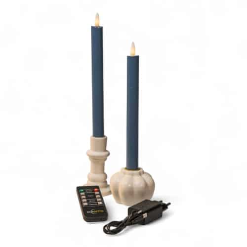 Twee blauwe LED-kaarsen in witte houders met een afstandsbediening en een zwarte voedingsadapter ervoor geplaatst, alles op een witte achtergrond.
