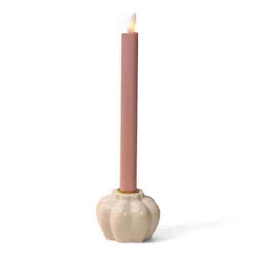 Een lange, roze conische kaars wordt aangestoken en in een glanzende, crèmekleurige kandelaar in de vorm van een pompoen geplaatst tegen een effen witte achtergrond.