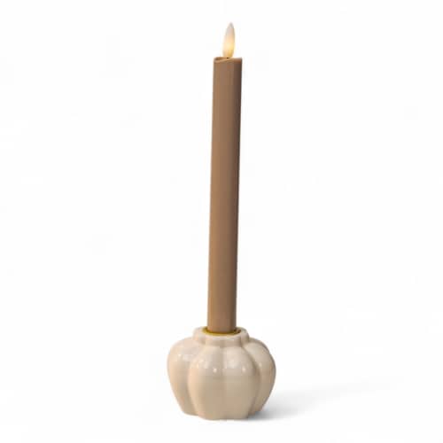 Een brandende beige conische kaars staat rechtop in een glanzende, crèmekleurige, pompoenvormige houder van keramiek tegen een effen witte achtergrond.