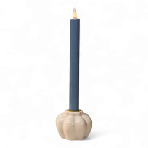 Een brandende donkerblauwe conische kaars staat in een ronde, crèmekleurige houder van keramiek met een geribbeld ontwerp, tegen een witte achtergrond.